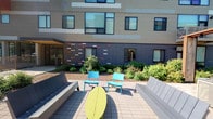 This outdoor courtyard is defined by its contemporary architectural backdrop and thoughtfully designed gathering space. The building facade combines contrasting materials, including a neutral upper section and a bold navy lower panel accented with colorful horizontal tile inlays in shades of red, white, and gray, creating a visually dynamic exterior. Large windows line the building at multiple levels, allowing natural light to flow both in and out of the adjacent interior spaces.

The courtyard itself is paved with neutral-toned interlocking pavers, providing a clean and durable surface underfoot. Seating is arranged in a sociable configuration, featuring bright teal accent chairs and a bold yellow-green oval table at the center, flanked by sleek, light-gray benches on either side. This mix of colors adds energy and personality to the outdoor space.

Lush landscaping borders the building's base, with layered plantings of shrubs, hostas, and other greenery that soften the transition between the hardscape and the structure. An entry door is visible to one side, framed by additional greenery. The courtyard receives generous natural sunlight, making it a welcoming and functional outdoor amenity well-suited for casual gatherings or quiet relaxation.
