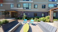This outdoor courtyard is defined by its contemporary architectural backdrop and thoughtfully designed gathering space. The building facade combines contrasting materials, including a neutral upper section and a bold navy lower panel accented with colorful horizontal tile inlays in shades of red, white, and gray, creating a visually dynamic exterior. Large windows line the building at multiple levels, allowing natural light to flow both in and out of the adjacent interior spaces.

The courtyard itself is paved with neutral-toned interlocking pavers, providing a clean and durable surface underfoot. Seating is arranged in a sociable configuration, featuring bright teal accent chairs and a bold yellow-green oval table at the center, flanked by sleek, light-gray benches on either side. This mix of colors adds energy and personality to the outdoor space.

Lush landscaping borders the building's base, with layered plantings of shrubs, hostas, and other greenery that soften the transition between the hardscape and the structure. An entry door is visible to one side, framed by additional greenery. The courtyard receives generous natural sunlight, making it a welcoming and functional outdoor amenity well-suited for casual gatherings or quiet relaxation.
