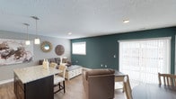 This single-floor space showcases a thoughtfully designed open-concept layout that integrates the living and dining areas into one cohesive, functional space. The standout feature is a bold teal accent wall in the living area, which adds a striking pop of color against the surrounding neutral gray walls, creating a visually dynamic yet balanced atmosphere.

The living area is comfortably sized, accommodating a sectional sofa, armchair, and a white storage coffee table. A small window and a set of vertical-blind-covered sliding doors allow natural light to filter into the space, brightening the interior. The light-toned laminate flooring runs throughout, contributing to a clean and contemporary aesthetic.

Transitioning seamlessly into the dining area, a dark-framed counter with a granite-style surface provides casual seating with upholstered bar-height chairs in neutral tones. Decorative wall mirrors with sunburst and medallion designs add an artistic touch to the space.

The textured popcorn ceiling with recessed lighting completes the interior, providing practical illumination. Overall, the space presents a modern, inviting environment with a cohesive blend of neutral tones and bold accents.