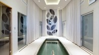 This sophisticated single-floor aquatic space is defined by its clean, modern aesthetic and carefully curated design elements. At its heart lies a pristine rectangular lap pool with calm, clear water, framed by polished light-toned flooring that reflects the space's overall sense of refinement. Floor-to-ceiling sheer white drapery lines both sides of the room, softening the architecture and contributing to a tranquil, spa-like ambiance. A dramatic focal point commands the far wall — a large-scale artistic installation composed of bold oval and circular forms rendered in a striking black-and-white palette, lending a contemporary gallery quality to the environment. The ceiling features a recessed tray design with integrated recessed lighting, providing a warm, even illumination throughout the space. Flanking the pool area, frosted glass panels marked with subtle signage indicate adjacent facilities, suggesting this space is part of a broader wellness or amenity complex. The overall design language is cohesive and intentional, balancing minimalist architecture with artistic expression to deliver an environment that is both functional and visually impressive — ideal for lap swimming or quiet relaxation.