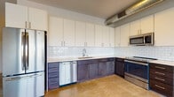 This single-floor, one-bedroom, one-bathroom unit showcases a thoughtfully designed industrial-modern aesthetic throughout. Polished concrete floors run continuously across the living and kitchen areas, complementing the unit's urban character. Exposed metallic ductwork runs along the ceiling, adding an authentic industrial touch that defines the overall design language.

The kitchen is a standout feature, equipped with stainless steel appliances including a refrigerator, dishwasher, range, and built-in microwave. Two-toned cabinetry pairs dark lower cabinets with crisp white upper cabinets, accented by a classic white subway tile backsplash and light countertops. The L-shaped layout provides ample workspace and storage.

The living area is generously proportioned, featuring large windows that flood the space with natural light and offer open views of the surrounding cityscape. A door from the living area appears to lead to an outdoor balcony or terrace, further enhancing the connection to the exterior.

The bathroom is clean and modern, featuring white cabinetry, a white countertop vanity with a large mirror, and a walk-in shower enclosed by black-framed glass doors with white subway tile surround. The hallway connecting the rooms maintains the same polished, minimal aesthetic found throughout the unit.