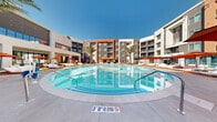 Located at 5050 W Russell Rd in Las Vegas, NV, this modern residential community is designed around a striking central courtyard that serves as the social heart of the property. The exterior architecture features a contemporary multi-story façade with a bold combination of stucco finishes in warm terracotta, charcoal gray, and crisp white, accented by sleek metal railings on private balconies throughout.

At the center of the community lies a generously sized resort-style pool with crystal-clear water, surrounded by a spacious deck furnished with wooden chaise lounges, red market umbrellas, and ample room for sunbathing beneath the Las Vegas sky. Tall palm trees frame the pool area, enhancing the resort atmosphere. Adjacent to the pool, covered outdoor terrace spaces feature large-format light tile flooring and a curated mix of modern seating arrangements, ideal for relaxing or socializing.

The ground-floor clubhouse is enclosed by floor-to-ceiling glass walls with sleek black metal framing, allowing abundant natural light to flood the interior while providing unobstructed views of the courtyard. Inside, the space is furnished with contemporary lounge seating in warm, neutral tones. Desert-inspired landscaping with decorative rock, native grasses, and drought-tolerant plantings lines the pathways connecting the various outdoor areas, reinforcing the property's harmonious integration with the Nevada environment.