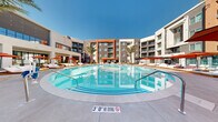 Located at 5050 W Russell Rd in Las Vegas, NV, this modern residential community is designed around a striking central courtyard that serves as the social heart of the property. The exterior architecture features a contemporary multi-story façade with a bold combination of stucco finishes in warm terracotta, charcoal gray, and crisp white, accented by sleek metal railings on private balconies throughout.

At the center of the community lies a generously sized resort-style pool with crystal-clear water, surrounded by a spacious deck furnished with wooden chaise lounges, red market umbrellas, and ample room for sunbathing beneath the Las Vegas sky. Tall palm trees frame the pool area, enhancing the resort atmosphere. Adjacent to the pool, covered outdoor terrace spaces feature large-format light tile flooring and a curated mix of modern seating arrangements, ideal for relaxing or socializing.

The ground-floor clubhouse is enclosed by floor-to-ceiling glass walls with sleek black metal framing, allowing abundant natural light to flood the interior while providing unobstructed views of the courtyard. Inside, the space is furnished with contemporary lounge seating in warm, neutral tones. Desert-inspired landscaping with decorative rock, native grasses, and drought-tolerant plantings lines the pathways connecting the various outdoor areas, reinforcing the property's harmonious integration with the Nevada environment.