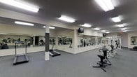 The Georgetown Oaks Fitness Center is a thoughtfully designed, single-floor exercise facility built to support a well-rounded workout routine. The space features a generous open floor plan finished with durable, gray carpet flooring suited for high-traffic athletic use. Bright fluorescent lighting mounted across the textured ceiling ensures the entire room is evenly and thoroughly illuminated throughout the day.

A defining architectural feature of the fitness center is the full-length mirror wall running along one side of the room, creating a sense of expanded space while allowing users to monitor their form during exercise. Structural columns with white trim detailing punctuate the floor plan, adding a subtle design element without interrupting the flow of the space.

The facility is equipped with a variety of cardio machines, including treadmills and a stationary bike, as well as cable-based strength training equipment, providing options for a comprehensive workout. Wall-mounted televisions are positioned throughout the room for entertainment during exercise sessions.

The overall ambiance is clean, functional, and well-maintained, reflecting a commitment to providing residents with a practical and accessible fitness amenity. The layout allows for comfortable movement between equipment, making the Georgetown Oaks Fitness Center a convenient and capable on-site wellness resource.