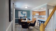 This two-floor residence offers a thoughtfully designed layout with a cohesive color palette of cool grays and rich teal accent walls that carry throughout the space, creating a sense of continuity and style. The open-concept dining and kitchen area serves as a central hub, where a warm-toned round dining table with sleek black velvet chairs sits beneath crown molding, adjacent to a fully equipped kitchen featuring honey-toned cabinetry, tile flooring, a full suite of white appliances including a range, dishwasher, and refrigerator, and a breakfast bar with additional seating. The living room is generously sized with plush carpeting, a bold teal feature wall adorned with framed artwork, and a sectional sofa centered around a marble-top coffee table. The primary bedroom is a serene retreat, highlighted by a teal accent wall, a large window with white drapes, and soft carpet underfoot. An additional bedroom offers neutral gray walls and comfortable proportions. Both bedrooms provide ample closet storage. The bathrooms are clean and functional, featuring warm wood vanities, tile flooring, and a tub-shower combination with white tile surround.