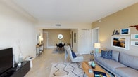 This single-floor residence offers an open, flowing layout that connects the living and dining areas into one cohesive space. The living area is anchored by a comfortable grey sofa accented with blue and patterned throw pillows, complemented by a chevron-upholstered accent chair and a wooden coffee table. A soft, textured area rug adds warmth underfoot, contrasting with the large-format light tile flooring that extends throughout the space.

Warm beige walls create a welcoming ambiance, while white ceilings with centrally placed HVAC vents contribute to a clean, finished look. The dining area, visible from the living room, features a compact round table paired with modern black chairs and is decorated with a round mirror, adding visual depth to the space.

A hallway near the entry leads to additional rooms, with a louvered closet door and a standard door visible along the wall, suggesting practical storage and private spaces beyond. A narrow console table near the entryway provides functional styling with decorative accessories. The overall aesthetic is casual yet curated, blending contemporary and coastal-inspired design elements throughout.