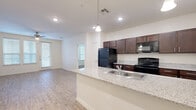 This single-floor residence offers a thoughtfully designed 2-bedroom, 2-bathroom layout that balances open communal spaces with private retreats. The heart of the home is an open-concept kitchen and living area, where light wood-look flooring flows seamlessly throughout. The kitchen features a peninsula with granite-look countertops, pendant lighting, rich dark-wood cabinetry, black appliances including a refrigerator, range, and microwave, and a stainless steel sink — all oriented toward the living area for an interactive, connected feel. The spacious living room is bathed in natural light from multiple windows fitted with blinds and is finished with a ceiling fan overhead. Both bedrooms are carpeted and well-sized, each offering ceiling fans and generous windows that invite natural light. The primary bedroom benefits from a dedicated en-suite bathroom, which includes dark-wood vanity cabinetry, a countertop sink, and direct access to a walk-in closet. The second bathroom mirrors this quality with similar finishes. Two closets, a hallway, and recessed lighting throughout contribute to the home's functional and clean aesthetic. Overall, the space presents a bright, neutral palette with consistent, modern finishes across every room.