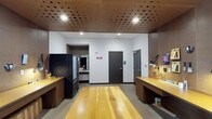 This single-floor space is defined by a striking architectural ceiling clad in perforated wood panels, lending warmth and acoustic character to the interior. Recessed lighting is integrated throughout the ceiling, providing even, focused illumination across the workspace. The flooring is a polished concrete surface, offering a clean and durable foundation that complements the industrial-yet-refined aesthetic of the space.

The main area features a large, solid wood central work table with a light natural finish, serving as a prominent focal point for collaborative or individual work. Flanking the central table on both sides are built-in wooden workbenches with storage cabinetry below, providing ample workspace and organization. Task lighting is mounted along the benches, supporting detailed work activities.

A black storage unit or equipment cabinet is positioned to one side, and a glass-doored refrigerator is visible in the background, indicating convenience amenities within the space. An adjoining area, accessible through an open passageway, appears to function as a utility or kitchenette zone. The space is enclosed by solid doors, ensuring privacy and separation from adjacent areas. White walls contrast with the warm wood tones throughout, creating a balanced and professional environment well-suited for workshop, studio, or technical workspace use.