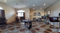 This single-floor space serves as a functional and inviting common area designed to accommodate a variety of activities. The room features a textured ceiling with recessed lighting, providing consistent, even illumination throughout. An arched window with decorative curtains allows natural light to filter in, adding warmth to the neutral, beige-toned walls.

The layout is thoughtfully divided into distinct zones. A central round dark-wood table paired with clear acrylic chairs serves as a casual gathering or dining area, anchored by a bold, circular-patterned carpet that adds visual interest underfoot. Adjacent to this, a pair of contemporary gray lounge chairs with a sleek chrome-accented side table creates a relaxed seating nook, complemented by tropical-themed wall art that lends a light, resort-inspired feel.

Toward the perimeter, a dedicated workspace includes a desk with office chairs, catering to those needing a quiet area for computer work or study. A tall bar-height table with a stool near a workstation further enhances the multi-functional nature of the space. Overall, the room balances practicality with comfort in a well-organized, approachable setting.