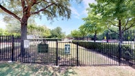 The Bark Park is a generously sized, fully enclosed outdoor space designed to provide a safe and enjoyable environment for dogs and their owners. Enclosed by sturdy black iron fencing, the park features a dedicated entry gate with posted rules and a welcoming sign, ensuring a secure space for pets to roam off-leash.

The grounds are divided into distinct zones, including an open grassy area where dogs can run freely and a paved concrete section bordered by a red-striped boundary, offering a firm surface suitable for active play. Several mature shade trees are distributed throughout the park, providing natural canopy coverage and relief from direct sunlight on warm days.

Rustic circular bench seating surrounds some of the larger trees, giving pet owners a comfortable place to rest while supervising their dogs. The park's open layout allows for clear sightlines across the entire space. Trimmed hedgerows along portions of the perimeter add a polished, well-maintained aesthetic. The overall ambiance is bright and welcoming, with abundant natural light and a relaxed, community-oriented atmosphere ideal for daily visits.
