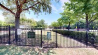 The Bark Park is a generously sized, fully enclosed outdoor space designed to provide a safe and enjoyable environment for dogs and their owners. Enclosed by sturdy black iron fencing, the park features a dedicated entry gate with posted rules and a welcoming sign, ensuring a secure space for pets to roam off-leash.

The grounds are divided into distinct zones, including an open grassy area where dogs can run freely and a paved concrete section bordered by a red-striped boundary, offering a firm surface suitable for active play. Several mature shade trees are distributed throughout the park, providing natural canopy coverage and relief from direct sunlight on warm days.

Rustic circular bench seating surrounds some of the larger trees, giving pet owners a comfortable place to rest while supervising their dogs. The park's open layout allows for clear sightlines across the entire space. Trimmed hedgerows along portions of the perimeter add a polished, well-maintained aesthetic. The overall ambiance is bright and welcoming, with abundant natural light and a relaxed, community-oriented atmosphere ideal for daily visits.