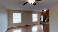 This single-story space showcases an open-concept layout that seamlessly connects the main living area to the kitchen. The floors throughout are finished in warm-toned hardwood, adding a polished and inviting quality to the space. A ceiling fan with an integrated light fixture is centrally mounted, providing both airflow and ambient lighting.

Two windows fitted with white horizontal blinds flank a white panel entry door along the front wall, allowing generous natural light to filter into the interior while maintaining privacy. The walls are painted in a neutral warm beige, contributing to a bright and clean aesthetic throughout.

The kitchen, visible to the right, is equipped with stainless steel appliances, including a French door refrigerator with a bottom freezer and ice dispenser. Warm wood-toned upper cabinetry complements the granite or stone-look countertops, lending a functional yet attractive finish to the cooking area.

Overall, the space presents a clean, practical design with consistent finishes and a light-filled atmosphere, well-suited for comfortable everyday living.