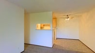 This single-floor space offers a clean, functional layout designed for comfortable everyday living. The main living area is carpeted in a neutral tone, providing a warm and understated foundation throughout the space. A defining architectural feature is the half-wall pass-through that connects the living area to the kitchen, creating an open sightline while maintaining a defined separation between spaces. The kitchen is outfitted with light wood cabinetry that adds a natural warmth to the room, and the layout appears efficient and well-organized. A ceiling fan with an integrated light fixture is centrally positioned in the living area, contributing to both air circulation and ambient lighting. The walls throughout are finished in a crisp, neutral white, offering a versatile backdrop suited to a variety of interior styles. A hallway extending from the main living space leads to additional rooms, suggesting a practical and straightforward floor plan. The overall ambiance of the space is bright, simple, and move-in ready, with a no-frills aesthetic that prioritizes function and adaptability.