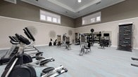 This expansive fitness center is thoughtfully designed to support a full range of workout routines across multiple dedicated zones. The main strength and conditioning area features elevated ceilings with clerestory windows that invite generous natural light, taupe-toned walls with crisp white wainscoting, and a geometric patterned carpet floor in soft grays and blues. The space is outfitted with a comprehensive selection of selectorized strength machines, a multi-functional cable and functional training station, a full dumbbell rack, and additional free weight equipment. Decorative wall art adds a polished aesthetic throughout. A dedicated cardio section houses a row of treadmills, each paired with wall-mounted flat-screen televisions for an engaging workout experience. The cycling studio offers a serene, spa-inspired atmosphere, anchored by a vibrant zen-themed wall mural, floor-to-ceiling mirrors, a large display screen, and a lineup of Spinning bikes alongside a rowing machine. An adjoining group exercise room provides an open, airy space with mirrored walls, high ceilings, and room for a variety of fitness classes. Together, these zones create a cohesive, professional-grade fitness environment designed for both performance and comfort.