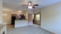 This single-floor space is designed with an open-concept layout that seamlessly connects the living area to the kitchen, creating a spacious and functional environment. The living area features neutral-toned carpeting underfoot and is equipped with a ceiling fan with an integrated light fixture, providing both comfort and illumination. A floor vent ensures efficient climate control throughout the space.

The kitchen is positioned adjacent to the living area and is defined by a raised breakfast bar with a dark countertop, offering a natural division between the two spaces while maintaining an open feel. Medium-toned wood cabinetry lines the kitchen walls, complemented by pendant lighting that adds focused illumination. The kitchen is equipped with black appliances, including a refrigerator and microwave, and is finished with matching dark countertops.

Beyond the main living and kitchen area, a hallway leads to additional rooms. A dedicated laundry room is visible, housing a washer and dryer and providing added convenience. Additional cabinetry and storage space can be seen further down the hallway. The overall ambiance is clean, neutral, and move-in ready, with a practical floor plan suited for everyday living.