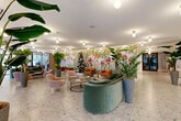 Located at 933 NW 12th St in Miami, FL, this single-floor lobby and corridor space offers a generously sized, thoughtfully designed environment that makes a memorable first impression. The main lobby is an expansive, open area anchored by a stunning full-wall botanical mural depicting lush tropical florals, perfectly suited to Miami's vibrant character. Polished terrazzo flooring runs throughout, lending a clean, timeless aesthetic that complements the warm, eclectic furnishings — including curved velvet sofas, blush pink armchairs, and cognac leather accent chairs arranged into inviting seating vignettes.

Tall potted tropical plants are strategically placed throughout, reinforcing the indoor-garden atmosphere. Globe pendant and flush-mount ceiling fixtures in brushed gold provide warm, ambient lighting across the space. A secondary lounge area features framed artwork, a large display screen, and additional curated seating, while a dedicated workstation counter with built-in mailboxes serves functional needs.

The hallway corridor connects the main spaces with elegance, lined with warm wood-paneled doors, olive-toned accent walls, decorative sconces, and sculptural gold-framed mirror panels that serve as artistic room dividers. Floor-to-ceiling sheer curtains along the glazed entry facade allow abundant natural light to filter in, enhancing the bright, airy ambiance throughout.