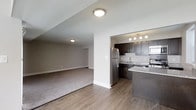 This single-floor residence showcases an open-concept design that seamlessly connects the kitchen and living areas. The kitchen is well-appointed with dark espresso cabinetry, granite countertops, and stainless steel appliances, including a full-size refrigerator, microwave, and range. A peninsula-style counter extends into the main living space, providing additional prep area and casual seating opportunities. Track lighting above the kitchen ensures ample task illumination. The flooring transitions from smooth hardwood-style planks in the kitchen and entry zone to plush, neutral-toned carpet in the living area, clearly delineating each functional space. White walls and flush-mount ceiling lighting keep the interior feeling bright and clean throughout. The living area is generously proportioned, offering flexible furniture arrangement options. The overall aesthetic is contemporary and practical, with a neutral color palette of whites, grays, and dark wood tones creating a cohesive and modern atmosphere suited for everyday living.