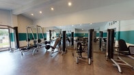 This single-floor fitness facility is thoughtfully designed to support a comprehensive workout experience across several distinct zones. The grand lobby entrance immediately sets the tone with soaring double-height ceilings, expansive floor-to-ceiling windows that flood the space with natural light, and rich hardwood flooring. A bold, graphic accent wall featuring colorful geometric shapes and a prominent reception desk anchors the welcoming area, while a statement chandelier adds a modern touch overhead.

The facility is organized into purposefully labeled zones. The cardio area, branded "balance," houses a row of treadmills, ellipticals, and steppers positioned along large windows that offer views of mature trees outside. The strength training room features professional-grade cable machines and weight benches, with full-length mirrors along the walls that visually expand the space and aid in form monitoring. A dedicated free weights corner, labeled "muscle," offers kettlebells, dumbbells, and a sturdy bench.

A connecting corridor labeled "energy" links the zones with polished concrete flooring and teal accent walls consistent throughout the facility. A private office or treatment room with yellow-painted walls and interior windows overlooking the gym floor provides staff with direct sightlines across the space. Recessed lighting throughout ensures consistent illumination across all areas.