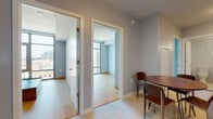 This contemporary urban residence is thoughtfully designed with clean lines, neutral tones, and high-quality finishes throughout. The open layout flows naturally between living, dining, and kitchen areas, with light hardwood flooring adding warmth to the cool grey palette of the walls. A compact dining area sits conveniently adjacent to the main corridor, anchored by a rounded wood table and modern chairs. The kitchen is well-appointed with white shaker-style cabinetry, stainless steel appliances including a gas range, a glossy white subway tile backsplash, and light stone countertops that create a crisp, functional workspace. The bedrooms are bright and airy, each benefiting from floor-to-ceiling windows that frame panoramic city views, flooding the rooms with natural light and offering a striking elevated perspective of the surrounding urban landscape. The bathrooms are finished to a high standard — one features a glass-enclosed walk-in shower with large-format grey wall tiles and a streamlined vanity, while the second offers a full soaking tub with a white tile surround, a generous vanity with an under-mount sink, and polished chrome fixtures. Throughout the space, the consistent use of light tones, modern hardware, and quality materials creates a cohesive, sophisticated aesthetic suited to contemporary urban living.