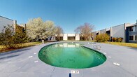 Located at 2444 E Maple Ave in Burlington, North Carolina, this outdoor space centers around a generously sized freeform pool set within a well-maintained concrete deck. The pool features a shallow entry point of approximately 2.4 feet and is flanked by metal entry rails for ease of access. The surrounding deck provides ample space for outdoor activities and relaxation.

The property is framed by a cluster of multi-story buildings with a modern, commercial aesthetic characterized by clean stucco and panel exteriors, blue accent trim, and large windows that overlook the outdoor grounds. The buildings form a courtyard-like enclosure around the pool area, creating a semi-private outdoor environment.

Mature deciduous and ornamental flowering trees are strategically placed throughout the grounds, adding natural character and seasonal color to the landscape. Manicured green lawns, pathway lighting, and decorative plantings further enhance the outdoor ambiance. Wide paved walkways connect the pool area to the surrounding buildings, offering convenient access throughout the complex.

The space benefits from open sky exposure, allowing for abundant natural light throughout the day. The combination of the pool, expansive deck, lush landscaping, and surrounding architecture creates a functional and visually cohesive outdoor environment suited for both leisure and gathering.