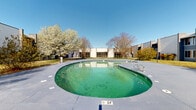 Located at 2444 E Maple Ave in Burlington, North Carolina, this outdoor space centers around a generously sized freeform pool set within a well-maintained concrete deck. The pool features a shallow entry point of approximately 2.4 feet and is flanked by metal entry rails for ease of access. The surrounding deck provides ample space for outdoor activities and relaxation.

The property is framed by a cluster of multi-story buildings with a modern, commercial aesthetic characterized by clean stucco and panel exteriors, blue accent trim, and large windows that overlook the outdoor grounds. The buildings form a courtyard-like enclosure around the pool area, creating a semi-private outdoor environment.

Mature deciduous and ornamental flowering trees are strategically placed throughout the grounds, adding natural character and seasonal color to the landscape. Manicured green lawns, pathway lighting, and decorative plantings further enhance the outdoor ambiance. Wide paved walkways connect the pool area to the surrounding buildings, offering convenient access throughout the complex.

The space benefits from open sky exposure, allowing for abundant natural light throughout the day. The combination of the pool, expansive deck, lush landscaping, and surrounding architecture creates a functional and visually cohesive outdoor environment suited for both leisure and gathering.
