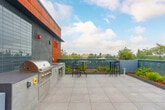 Located atop 2650 Telegraph Ave in Berkeley, CA, this expansive rooftop terrace offers a well-appointed outdoor amenity space with sweeping views of the Berkeley Hills, the surrounding urban canopy, and distant bay vistas. The terrace is finished with large-format light gray porcelain tile throughout, creating a clean, contemporary aesthetic that complements the building's modern exterior of paneled metal cladding and warm weathered steel accents.

At the center of the space sits a substantial fire pit encased in a glass wind screen, flanked by sleek black bar-height tables and stools — ideal for gathering and entertaining. Along the building's rear facade, a fully equipped outdoor kitchen is built into a poured concrete counter, featuring a stainless steel multi-burner gas grill and a utility sink with ample cabinet storage below. The facade itself combines textured tile, corten-style panels, and smooth stucco, adding architectural character to the setting.

Lush raised planters line the perimeter, filled with ornamental grasses, shrubs, and slender trees that provide greenery and a sense of enclosure without obstructing the open sky views. The transparent glass perimeter railing preserves unobstructed sightlines across Berkeley's rooftops and hillside. This thoughtfully designed rooftop terrace functions as a dynamic communal outdoor space suited for both relaxed gatherings and social events.