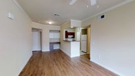 This single-floor space is designed with an open-concept layout that seamlessly connects the main living area to the kitchen. Warm-toned hardwood flooring runs throughout, lending a cohesive and inviting feel to the interior.

The living area is bright and spacious, accented by crown molding along the ceiling and a ceiling fan with integrated lighting. A built-in desk nook with open shelving is tucked along one wall, offering a practical and organized workspace without occupying additional floor space.

The kitchen is defined by a white peninsula that separates it from the living area while maintaining visual openness. Rich, dark-toned cabinetry provides a striking contrast against the lighter walls and appliances, which include a white refrigerator and range. A recessed light above the kitchen adds focused illumination to the space.

Doorways visible from the main area lead to additional rooms, suggesting a well-considered floor plan. The neutral wall color, combined with the warm flooring and classic crown molding details, gives the space a clean, timeless aesthetic suitable for a variety of furnishing styles.