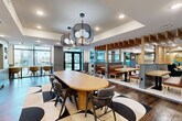 Located at 3305 South Blvd in Charlotte, NC, this thoughtfully designed single-floor amenity space offers a dynamic blend of social and functional zones across a generous open layout. The space is anchored by a central dining area featuring a large oval wood table surrounded by upholstered chairs, set atop a bold geometric area rug and illuminated by striking sculptural pendant lighting. Tray ceilings add architectural dimension throughout.

The kitchen area runs along one wall with sleek gray cabinetry, stainless steel appliances, white quartz countertops, and a distinctive illustrated backsplash. Open wood shelving above displays lush potted plants, adding a fresh, organic touch. Floor-to-ceiling windows at one end flood the space with natural light and provide views of an outdoor terrace.

A cozy lounge zone anchors another section of the space, featuring an exposed brick accent wall adorned with vibrant abstract artwork, a wall-mounted television framed by warm wood slat paneling, and plush velvet swivel chairs arranged around a coffee table — ideal for relaxed gathering.

Additional seating is provided through banquette-style booths with teal upholstery and wood accents, separated by open shelving partitions. Rich hardwood flooring flows throughout, tying together the various areas into a cohesive, design-forward community space.