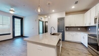 Located at 2300 Laurel Rd E in Nokomis, Florida, this single-floor residence showcases a clean, modern design with thoughtfully selected finishes throughout. The open-concept kitchen serves as the heart of the space, anchored by a large center island with a quartz countertop, an undermount sink, and elegant globe pendant lights overhead. Crisp white shaker-style cabinetry lines the walls, complemented by a gray subway tile backsplash and a full suite of stainless steel appliances, including a range, microwave, and refrigerator. Wood-look luxury vinyl plank flooring runs throughout the main living areas, providing warmth and continuity. The bedroom features plush carpeting and neutral tones, offering a comfortable retreat. The bathroom is finished with wood-look flooring, a glass-enclosed walk-in shower with tile surround, and gray-paneled doors that echo the design language found throughout the unit. Recessed lighting and contemporary fixtures provide consistent, well-distributed illumination in every room. The overall aesthetic is cohesive and modern, combining light walls, gray accent doors, and quality materials to create a fresh, move-in-ready living environment.