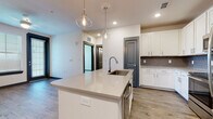 Located at 2300 Laurel Rd E in Nokomis, Florida, this single-floor residence showcases a clean, modern design with thoughtfully selected finishes throughout. The open-concept kitchen serves as the heart of the space, anchored by a large center island with a quartz countertop, an undermount sink, and elegant globe pendant lights overhead. Crisp white shaker-style cabinetry lines the walls, complemented by a gray subway tile backsplash and a full suite of stainless steel appliances, including a range, microwave, and refrigerator. Wood-look luxury vinyl plank flooring runs throughout the main living areas, providing warmth and continuity. The bedroom features plush carpeting and neutral tones, offering a comfortable retreat. The bathroom is finished with wood-look flooring, a glass-enclosed walk-in shower with tile surround, and gray-paneled doors that echo the design language found throughout the unit. Recessed lighting and contemporary fixtures provide consistent, well-distributed illumination in every room. The overall aesthetic is cohesive and modern, combining light walls, gray accent doors, and quality materials to create a fresh, move-in-ready living environment.