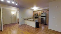 This single-floor residence centers around an open-concept kitchen and living area designed for both functionality and comfort. The kitchen is well-appointed with stainless steel appliances, including a refrigerator, electric range, and over-the-range microwave. Warm-toned wood cabinetry provides ample storage above and below the countertops, which feature granite surfaces that extend across the peninsula. The peninsula itself offers casual seating with built-in shelving underneath, making it a practical space for dining or food preparation. A single-basin sink with a contemporary arc faucet is seamlessly integrated into the peninsula countertop. The hardwood flooring throughout the main living space adds warmth and continuity, reflecting the ambient and track lighting overhead. A hallway visible from the main area leads to additional rooms, including what appears to be a bathroom with matching cabinetry. The neutral wall tones and consistent finishes throughout create a cohesive, understated aesthetic that feels clean and inviting. Overall, the layout is efficient and practical, making good use of the available space.