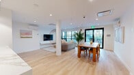 Located at 750 Robinson Street in Coquitlam, BC, this thoughtfully designed single-floor amenity space seamlessly integrates a living room, dining area, and kitchen into one cohesive open-plan layout. Light oak hardwood flooring flows throughout, anchoring the bright, modern aesthetic defined by crisp white walls and recessed lighting.

The living area is a standout feature, centered around a striking wood-slatted accent wall with an arched detail framing a wall-mounted television. A plush sectional sofa, accent armchair, and a low-profile coffee table create an inviting lounge setting, while floor-to-ceiling windows flood the space with natural light and offer views of the surrounding neighbourhood.

The dining area comfortably accommodates a full-sized wooden dining table and chairs, positioned adjacent to black-framed glass doors that open directly to an outdoor terrace. Curated artwork adorns the walls, adding a refined, gallery-like quality to the room.

The kitchen is elegantly appointed with warm amber cabinetry, a dramatic marble-look slab backsplash, white quartz countertops, and a peninsula bar with seating for three. Integrated stainless steel appliances, including a refrigerator and microwave, complete the functional yet stylish culinary space. The overall ambiance is modern, welcoming, and well-appointed.