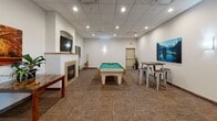 Located at 2915 Bluestem Dr in West Fargo, North Dakota, this single-floor community game room offers a well-appointed and inviting recreational space designed for socializing and leisure. The centerpiece of the room is a full-size billiards table set atop textured, neutral-toned carpet that extends throughout the space, providing a comfortable and stylish foundation. A built-in fireplace with a tile surround anchors one wall, topped by a wall-mounted flat-screen television set within an arched alcove — creating a cozy focal point ideal for gathering. A dedicated cue rack keeps billiards equipment neatly organized nearby. The room is accented with vibrant canvas artwork throughout, including nature-inspired landscape pieces that add color and character to the crisp white walls. Recessed lighting in the drop ceiling provides bright, even illumination across the space, complemented by elegant wall sconces in the adjoining corridor. High-top tables with industrial-style stools offer additional seating for guests. The corridor connecting to the main room is tastefully decorated with bold abstract artwork and wall-mounted lighting, creating a polished transition between areas. Overall, the space strikes a balance between functional recreation and refined communal design, making it an attractive amenity for residents and visitors alike.