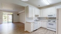 This single-floor residence offers a clean, functional layout designed for comfortable everyday living. The open-concept kitchen flows seamlessly into the living area, creating a spacious and connected feel throughout the main living space. The kitchen is well-appointed with white cabinetry offering generous storage, light-toned countertops, and a full suite of white appliances including a refrigerator, electric range, built-in microwave, and dishwasher. A pendant light fixture with a circular skylight detail overhead adds a subtle architectural touch to the kitchen area.

Light wood-look flooring runs throughout the kitchen and living areas, contributing to a bright and airy atmosphere. Large sliding glass doors in the living room flood the space with natural light and provide views of the surrounding greenery. The bedroom features plush carpeting and a broad window that frames pleasant outdoor views, creating a calm and restful environment.

The bathroom includes a dual-sink vanity with white cabinetry, a large wall mirror with overhead lighting, and tile flooring. A dedicated laundry closet houses a top-load washer with shelving above for added storage convenience. Overall, the space presents a cohesive, light-filled interior with a neutral palette throughout.