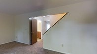 This two-floor space is characterized by its clean, neutral aesthetic and functional layout. The upper level, visible in the image, features wall-to-wall carpeting in a warm beige tone that adds a sense of comfort underfoot. A prominent staircase with a diagonal railing transitions between floors, its angular design creating a striking architectural detail against the white walls.

Adjacent to the staircase landing is a doorway that opens into a bathroom, which is partially visible and features warm wood-toned cabinetry providing ample storage. A window in the bathroom allows natural light to filter into the space, brightening the room. Recessed lighting in the hallway area supplements the natural light, ensuring the transition areas remain well-lit.

The walls throughout are finished in a crisp, neutral white, contributing to a bright and open atmosphere. The consistent use of neutral tones across the flooring and walls creates a cohesive and versatile backdrop. Overall, the space presents a straightforward, well-maintained interior with practical design choices suited to everyday living.