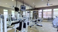 This fitness center offers a thorough and well-organized workout facility designed to accommodate a wide range of strength training and exercise needs. The space is outfitted with an extensive array of cable machines, weight machines, and multi-functional strength training equipment, all finished in a clean black-and-white aesthetic. Ceiling-mounted televisions are positioned throughout the room, ensuring users can stay entertained during their workouts. Full-length mirrored walls line one side of the facility, visually expanding the space and allowing for proper form monitoring during exercise. Large windows with horizontal blinds are installed along the perimeter walls, allowing ample natural light to fill the room and providing views of the surrounding greenery. The elevated ceilings contribute to an open, airy atmosphere that prevents the space from feeling cramped despite the density of equipment. The flooring features a neutral, multi-toned tile pattern that complements the contemporary design of the room. Recessed lighting supplements the natural light, ensuring consistent illumination throughout all hours. The overall ambiance is clean, modern, and purpose-driven, making this fitness center a well-appointed destination for comprehensive strength and conditioning training.