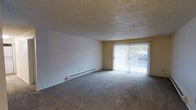 This single-floor space offers a straightforward and functional layout designed for comfortable everyday living. The main living area is finished with wall-to-wall carpeting that adds warmth underfoot, while a textured popcorn ceiling runs throughout, giving the space a classic residential character. Smooth, neutral-toned walls provide a clean backdrop that lends itself well to a variety of furnishing styles. A prominent sliding glass door serves as a key architectural feature, fitted with vertical blinds and allowing natural light to filter into the room while offering a direct connection to the outdoors. The view through the glass door hints at greenery beyond, adding a pleasant natural element to the interior ambiance. A baseboard heater runs along the lower portion of one wall, providing an efficient and unobtrusive heating solution. The open nature of the living area creates a sense of spaciousness, making it versatile for various living arrangements. Overall, the space presents a clean, unpretentious environment that prioritizes practicality and ease of use, with the sliding glass door and natural light standing out as its most notable features.