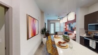 This single-floor residence showcases a thoughtfully designed open-concept layout that connects the kitchen, dining, and living areas into one cohesive space. The kitchen is equipped with stainless steel appliances—including a refrigerator, range, microwave, and dishwasher—and features rich dark cabinetry extending to the ceiling, a tile backsplash, and a generous island with a quartz countertop that doubles as a casual dining surface. Pendant lighting illuminates the island, while recessed lighting brightens the full kitchen area. Wood-look flooring runs throughout the main living spaces, adding warmth and continuity. The living area benefits from natural light through large windows with blinds and a sliding glass door, complemented by a ceiling fan overhead. A bold accent wall provides a vibrant backdrop to the living space. The bedroom is distinguished by a deep teal accent wall and a ceiling fan, with large windows allowing ample natural light. The bathroom features dark cabinetry, granite countertops, an illuminated mirror, and wood-look flooring, with additional doors leading to closet and storage areas. Overall, the space presents a modern, colorful aesthetic with cohesive design elements throughout.