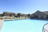 This outdoor recreational area centers around a large, inviting swimming pool with crystal-clear blue water, designed to serve as a premier amenity for the surrounding residential community. The pool is generously sized, offering ample room for both leisurely swimming and active use, with clearly marked depth indicators along its edges.

Surrounding the pool is a broad concrete sundeck that provides plenty of open space for relaxation and recreation. Multiple slatted lounge chairs are arranged along the deck, offering comfortable spots for sunbathing under the open sky. A patio dining set with chairs is also positioned poolside, creating a convenient area for outdoor gatherings and casual dining.

The entire pool area is enclosed by a sturdy wrought iron security fence with a gated entry, ensuring safety and controlled access. The facility is monitored by an ADT security system, as indicated by signage on the adjacent building. A single-story building with vinyl siding and a pitched roof sits directly adjacent to the pool, featuring large windows that overlook the water, and appears to serve as a clubhouse or amenity center with pool-facing access doors.

Mature trees and well-maintained landscaping are visible beyond the fencing, providing a natural backdrop and some shade. The space benefits from abundant natural light and open sky views, creating a bright and welcoming atmosphere throughout the day.