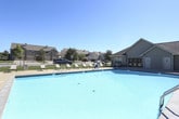 This outdoor recreational area centers around a large, inviting swimming pool with crystal-clear blue water, designed to serve as a premier amenity for the surrounding residential community. The pool is generously sized, offering ample room for both leisurely swimming and active use, with clearly marked depth indicators along its edges.

Surrounding the pool is a broad concrete sundeck that provides plenty of open space for relaxation and recreation. Multiple slatted lounge chairs are arranged along the deck, offering comfortable spots for sunbathing under the open sky. A patio dining set with chairs is also positioned poolside, creating a convenient area for outdoor gatherings and casual dining.

The entire pool area is enclosed by a sturdy wrought iron security fence with a gated entry, ensuring safety and controlled access. The facility is monitored by an ADT security system, as indicated by signage on the adjacent building. A single-story building with vinyl siding and a pitched roof sits directly adjacent to the pool, featuring large windows that overlook the water, and appears to serve as a clubhouse or amenity center with pool-facing access doors.

Mature trees and well-maintained landscaping are visible beyond the fencing, providing a natural backdrop and some shade. The space benefits from abundant natural light and open sky views, creating a bright and welcoming atmosphere throughout the day.
