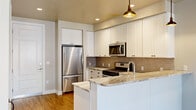 This efficiently designed single-floor unit offers a well-appointed layout comprising a kitchen, living room, and bathroom, making smart use of every square foot. The kitchen is a standout feature, showcasing white shaker cabinetry reaching toward the ceiling, stone countertops with striking veining, a classic subway tile backsplash, and a full suite of stainless steel appliances including a gas range, built-in microwave, dishwasher, and refrigerator. A farmhouse-style stainless sink and a peninsula with bar-height counter space add both function and character, while industrial-style pendant lights lend a modern touch. Warm hardwood-style flooring flows seamlessly throughout the space, complementing the neutral wall tones. The living area is bathed in natural light through large floor-to-ceiling windows fitted with horizontal blinds, offering views of the surrounding urban landscape. A sliding barn door adds a stylish architectural detail. A stacked washer and dryer unit is neatly integrated into the space. The bathroom continues the upscale aesthetic with a marble-veined stone vanity countertop, an undermount sink, and a boldly framed mirror, delivering a polished, cohesive finish to the overall design.
