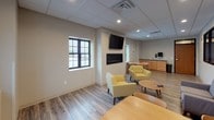 This single-floor communal space is thoughtfully designed to provide a comfortable and functional environment for relaxation and social interaction. The open layout features light wood-look flooring throughout, neutral wall tones, and a drop tile ceiling with recessed lighting that creates a bright, welcoming ambiance.

The lounge area serves as the focal point of the space, anchored by a sleek linear fireplace topped with a wall-mounted flat-screen television. Comfortable seating in warm yellow upholstered armchairs and a grey loveseat are arranged around a round wooden coffee table, encouraging casual conversation and relaxation. An interior window with black-framed mullions offers a view into an adjacent fitness or activity area, adding depth and connectivity to the overall space.

A dining area accommodates multiple occupants at a wooden table with coordinating chairs, ideal for shared meals or group gatherings. Along the back wall, a practical kitchenette is equipped with warm wood cabinetry, a dark countertop, a sink, and a paper towel dispenser, providing convenient amenities for everyday use. Natural light enters the space through black-framed exterior windows, complementing the clean, contemporary aesthetic throughout. The overall design strikes a balance between functional utility and inviting comfort.