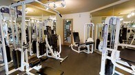 This fitness center offers a focused and functional workout environment designed to accommodate a range of strength training needs. The space is outfitted with multiple professional-grade cable and weight machines, including a multi-function cable station, a leg extension/curl machine, and additional resistance equipment — all finished in a clean white-and-black color scheme. Rubber flooring throughout provides durability and safety underfoot, making the space practical for active use. Floor-to-ceiling mirrors line the walls, visually expanding the room and allowing users to monitor their form during exercise. Overhead, adjustable track lighting illuminates the space evenly, ensuring excellent visibility across all workout stations. A windowed door at the rear of the room introduces a hint of natural light and provides access to an adjoining area. The overall ambiance is clean, organized, and purpose-driven, reflecting a well-maintained facility suited for consistent, dedicated use.