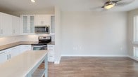 This single-floor residence offers a thoughtfully designed open-concept layout that seamlessly connects the living area and kitchen. Rich hardwood-style flooring extends throughout the main living space, complementing the neutral, light-toned walls and creating a warm yet contemporary atmosphere. Large floor-to-ceiling windows flood the living area with abundant natural light while providing views of the surrounding urban environment. A ceiling fan adds both functionality and a finishing touch to the spacious living room.

The kitchen is well-equipped and modern, featuring white shaker-style cabinetry with brushed nickel hardware, dark quartz countertops, and a white subway tile backsplash. Stainless steel appliances — including a French door refrigerator with ice dispenser, electric range, over-the-range microwave, and dishwasher — provide a complete culinary setup. A kitchen island with a light countertop offers additional workspace and casual dining potential.

The bathroom is finished with consistent design details, including white cabinetry, a dark quartz vanity top, an undermount sink, a large framed mirror, and a tiled shower-tub combination with chrome fixtures. The overall design palette is clean, cohesive, and modern throughout.