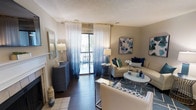 This well-appointed single-floor residence offers an open-concept layout that seamlessly connects the living, dining, and kitchen areas. The living room serves as the centerpiece of the space, anchored by a classic white-trimmed fireplace with a tile surround and a wall-mounted flat-screen television above. Rich, dark hardwood flooring runs throughout, providing a striking contrast against the warm neutral-toned walls. Natural light pours in through a sliding glass door that opens to a private balcony, framed by elegant ombré navy-to-white sheer curtains that add a refined, decorative touch. The living area is thoughtfully furnished with cream upholstered seating, a geometric-patterned area rug in blue and white, and coordinating navy accent pieces including a decorative chest and stylish side tables. The dining area, positioned adjacent to the kitchen, features a round table with dark chairs beneath a sunburst mirror accent. The kitchen opens to the main living space and showcases light marble-style countertops. Recessed lighting and textured ceilings complete the interior, while a hallway leads to additional private rooms, making this a comfortable and cohesive living environment with a polished coastal-inspired aesthetic.