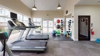 This single-floor fitness center is thoughtfully designed to support a comprehensive workout experience. The main exercise area is bright and airy, featuring large floor-to-ceiling windows and glass double doors that flood the space with natural light and offer views of the surrounding exterior. Vaulted ceilings with pendant lighting add a sense of openness and volume to the room.

The cardio zone is well-appointed with multiple treadmills and elliptical machines, each equipped with individual screens. Full-length mirrors line one wall, enhancing the sense of space while allowing users to monitor their form. A dedicated functional training area houses a colorful collection of medicine balls, stability balls, and resistance equipment arranged on protective floor mats.

The free weights section features a tiered dumbbell rack with a wide range of weights, accompanied by a water cooler, exercise mats, and a resistance band station. Motivational fitness-themed artwork and wall graphics add energy and character to the space throughout.

A hallway connects to an accessible restroom, finished with tile flooring and safety grab bars. The overall design maintains a clean, neutral palette with warm tones, creating a welcoming and functional environment suited for a variety of fitness routines.