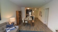 This single-floor residence showcases a thoughtfully designed open-concept layout that seamlessly connects the living, dining, and kitchen areas. Hardwood-style flooring in a rich, dark tone runs throughout the main living spaces, lending warmth and visual continuity to the interior.

The dining area is anchored by a wooden table paired with upholstered chairs and a matching bench, creating a casual yet refined setting. Adjacent to it, the kitchen features dark cabinetry, stainless steel appliances including a full-sized refrigerator, and a peninsula with a sink that doubles as a breakfast bar. Pendant lighting above the peninsula adds a modern touch, complementing the flush-mount ceiling fixtures that illuminate the space.

A bookcase with warm wood tones is positioned near the living area, providing functional storage and a decorative accent. The living area features a plush area rug and a sofa, creating a comfortable retreat. The hallway leads to the entry door, with framed wall art adding character to the corridor. The overall ambiance is contemporary and cohesive, with a neutral color palette that enhances the sense of openness.