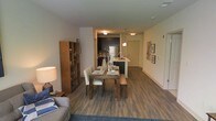 This single-floor residence showcases a thoughtfully designed open-concept layout that seamlessly connects the living, dining, and kitchen areas. Hardwood-style flooring in a rich, dark tone runs throughout the main living spaces, lending warmth and visual continuity to the interior.

The dining area is anchored by a wooden table paired with upholstered chairs and a matching bench, creating a casual yet refined setting. Adjacent to it, the kitchen features dark cabinetry, stainless steel appliances including a full-sized refrigerator, and a peninsula with a sink that doubles as a breakfast bar. Pendant lighting above the peninsula adds a modern touch, complementing the flush-mount ceiling fixtures that illuminate the space.

A bookcase with warm wood tones is positioned near the living area, providing functional storage and a decorative accent. The living area features a plush area rug and a sofa, creating a comfortable retreat. The hallway leads to the entry door, with framed wall art adding character to the corridor. The overall ambiance is contemporary and cohesive, with a neutral color palette that enhances the sense of openness.