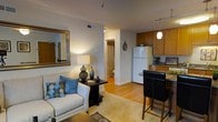 This single-floor residence offers a thoughtfully designed open-concept layout that seamlessly connects the living area to the kitchen, creating a sense of spaciousness and flow. The living space features warm hardwood-style flooring, neutral-toned walls, and a cozy seating area accented with decorative throw pillows and a stylish side table. A large framed mirror adds depth and reflects natural and ambient light throughout the room. Track lighting overhead provides additional illumination, complementing the warm glow of the table lamp. The kitchen is well-equipped with warm-toned wooden cabinetry, granite-style countertops, and a full-size refrigerator. A breakfast bar with dark leather seating offers a casual dining option, while a pendant light above adds a modern touch. The open sightlines between the kitchen and living area make the space feel cohesive and functional. A hallway visible from the main living area leads to a bathroom, complete with a curtained shower and toilet, maintaining a clean and practical design. Overall, the space strikes a balance between comfort and functionality, making it an inviting and efficient home.