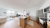 This single-floor residence is designed with a contemporary, minimalist sensibility, featuring warm light wood flooring and crisp white walls that create a bright, cohesive atmosphere throughout. The open-concept kitchen and living area serves as the heart of the space, where a central island with warm wood cabinetry and a white quartz countertop anchors the layout. The island is equipped with a sink, dishwasher, and seating for casual dining, while stainless steel appliances—including a full-size refrigerator—complete the functional kitchen setup. Pendant lighting above the island adds a modern design touch.

The living area flows seamlessly from the kitchen, offering generous space bathed in natural light from well-placed windows that provide outdoor views. The bedroom features light wood flooring, ample wall space, and access to a dedicated closet fitted with sliding doors that maximize usability.

The unit includes two bathrooms. One features dark tile flooring, a frameless sliding glass shower enclosure, and clean white finishes for a sleek, modern look. The second bathroom offers a shower-tub combination with a curtain, dark tile flooring, and a wood-accented vanity. Both bathrooms are finished with contemporary fixtures throughout.